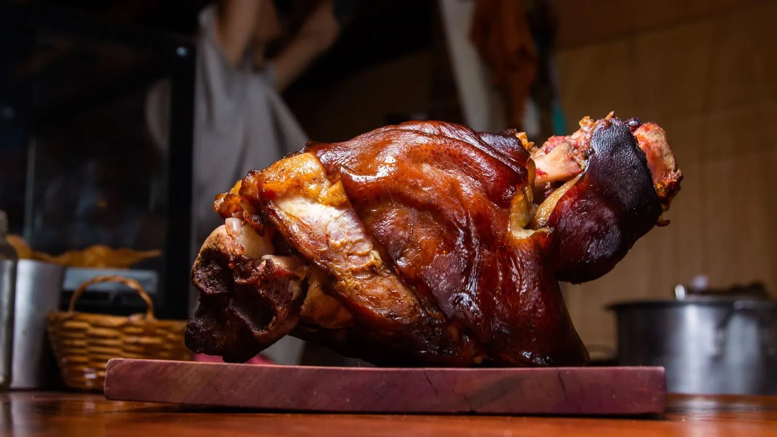 Codillo de cerdo entero asado sobre una tabla de madera, con la piel brillante de tono caoba y el hueso asomando por los extremos, ante una cocina desenfocada