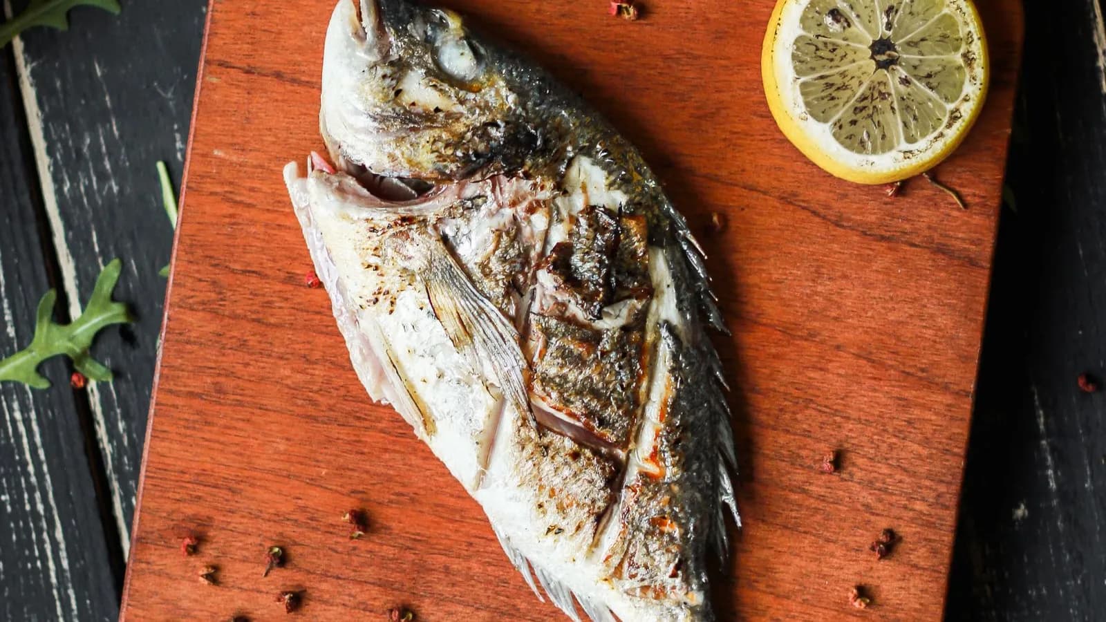 Dorada entera a la brasa con un limón asado y granos de pimienta sobre una tabla de madera oscura