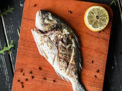 Dorada entera a la brasa con un limón asado y granos de pimienta sobre una tabla de madera oscura