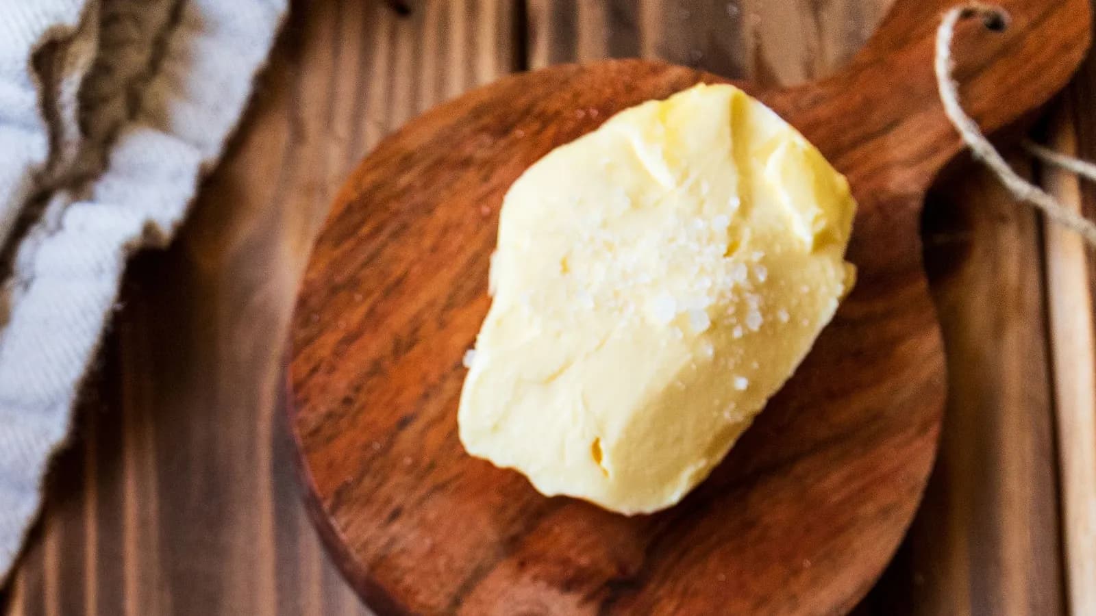 Porción de mantequilla cremosa y dorada con escamas de sal gruesa por encima, sobre una pequeña tabla de madera redonda con mango y cordel, en una mesa de tablones oscuros junto a un paño blanco