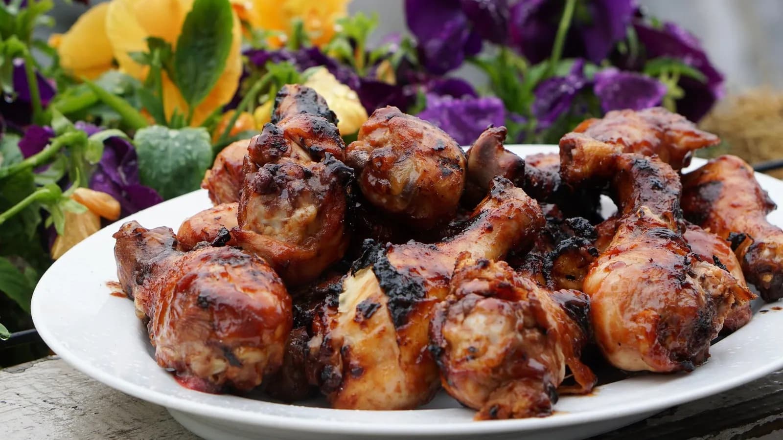Montón de muslos de pollo con un glaseado oscuro y brillante y zonas churruscadas, servidos en un plato blanco sobre madera envejecida, con flores amarillas y moradas desenfocadas al fondo