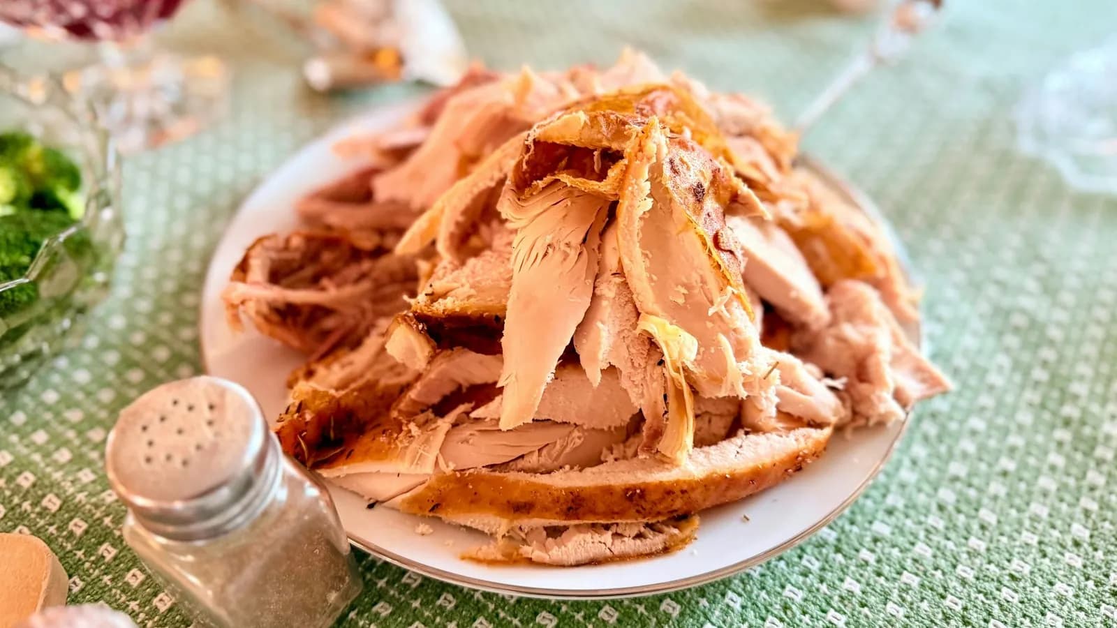 Plato blanco con un montón de pavo asado en trozos y lonchas, carne blanca con la piel dorada, sobre un mantel verde; en primer plano un salero y un pimentero de cristal y, desenfocados, cuencos con salsa de arándanos y brócoli