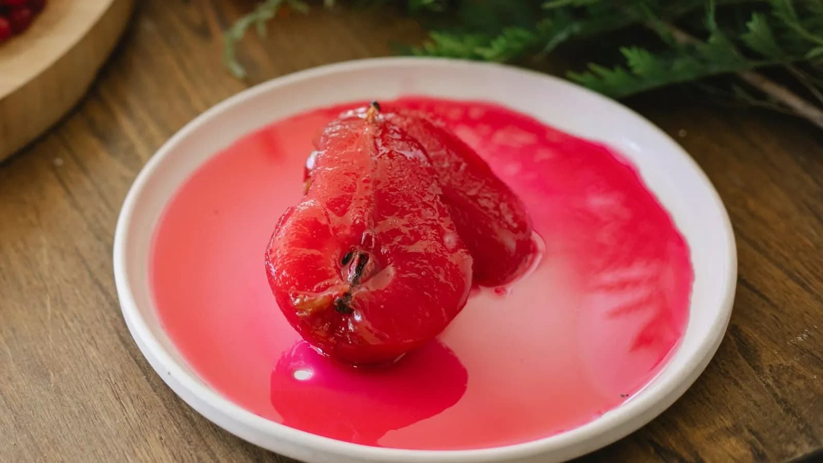 Una pera pochada partida en dos mitades, de un rojo intenso y brillante, en almíbar rosado sobre un plato blanco; al fondo, un bizcocho con grosellas y ramas de abeto