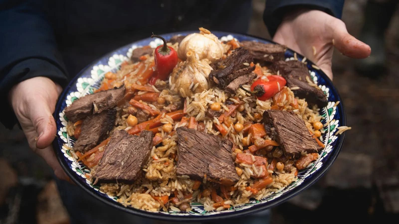 Plov uzbeko servido en una fuente de cerámica con el borde decorado en azul, blanco y verde que una persona sostiene con las dos manos al aire libre: arroz suelto con tiras gruesas de zanahoria y garbanzos, grandes lonchas de carne por encima, dos cabezas de ajo enteras y dos guindillas rojas (versión con garbanzos)
