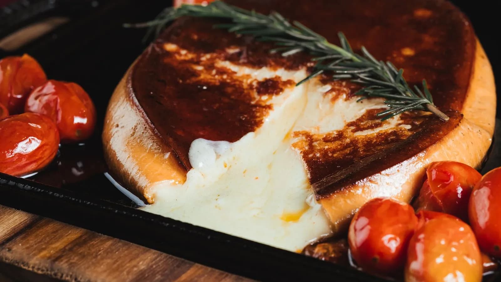 Rueda de provolone fundido a la brasa en sartén de hierro fundido con tomates cherry asados y una ramita de romero