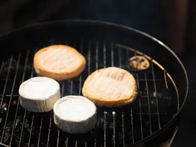 Four round cheeses smoking on the grate of a black barbecue with faint wisps of smoke