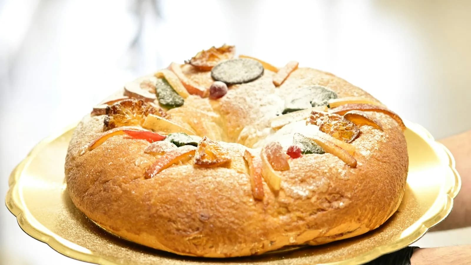 Rosca de Reyes de estilo mexicano entera, sobre una bandeja dorada de borde festoneado que sostienen unas manos, una con guante negro: corona de masa dorada espolvoreada de azúcar glas y cubierta de fruta confitada de colores (rodajas caramelizadas, tiras anaranjadas y piezas verdes, rojas y oscuras), ante un fondo claro desenfocado