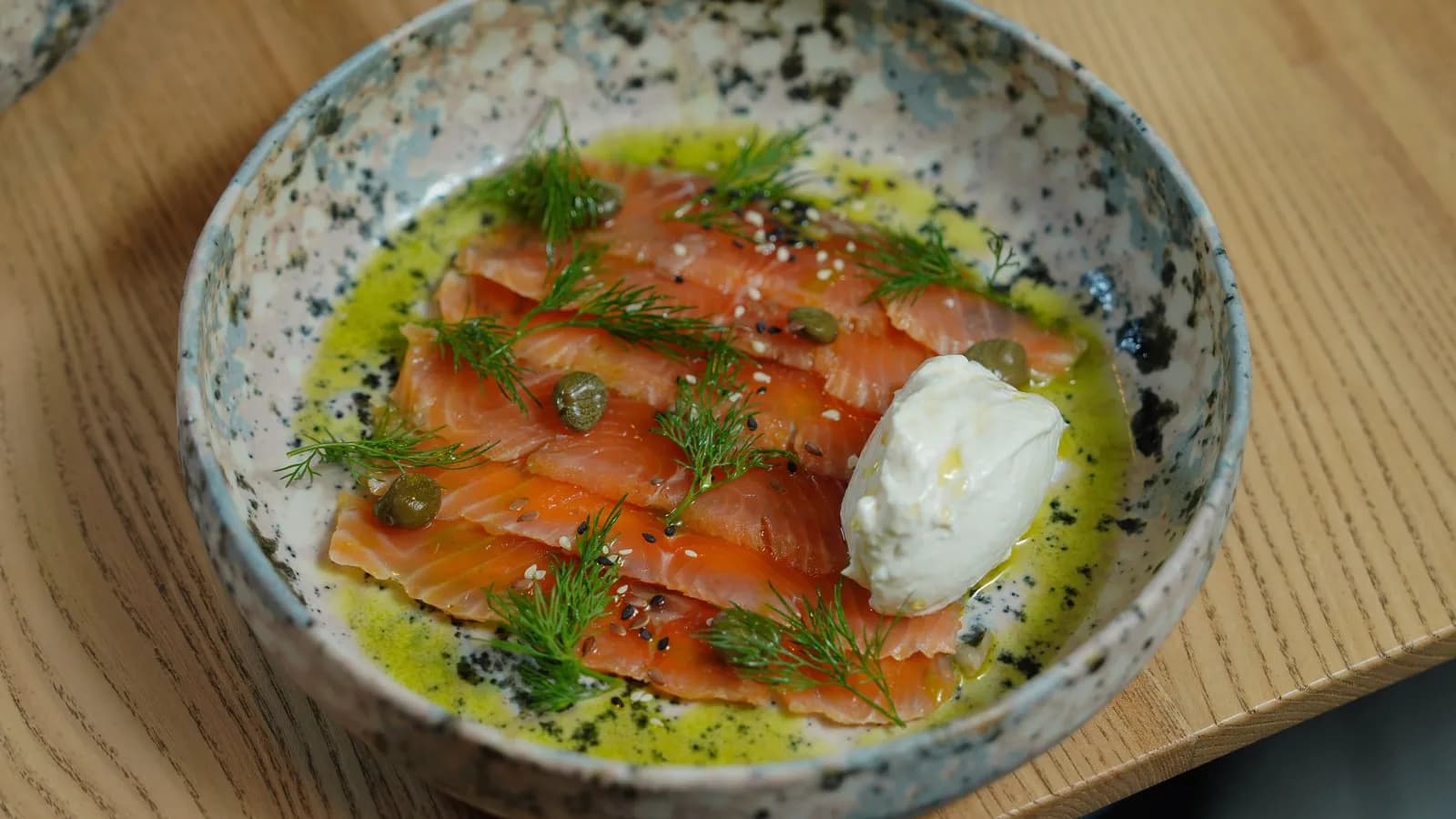 Lonchas finas de salmón ahumado con eneldo, alcaparras y sésamo en un cuenco de cerámica moteada, con aceite de oliva y una cucharada de crema blanca, sobre una mesa de madera clara