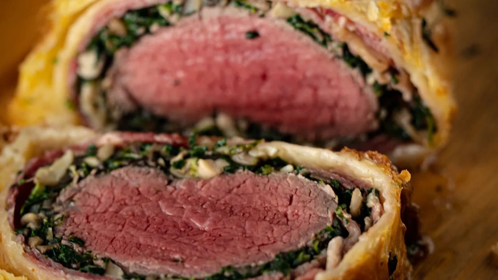Primer plano de un solomillo Wellington cortado: el centro de vacuno rosado, rodeado de una capa fina de jamón, otra de espinacas y champiñón picados y hojaldre dorado, con una rodaja separada delante sobre una tabla de madera