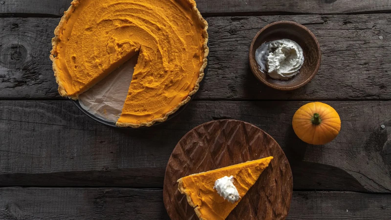 Tarta de calabaza vista desde arriba, de relleno naranja liso y borde de masa dorada, con una porción retirada; debajo, una porción con nata montada sobre una tabla de madera tallada, y a la derecha un cuenco de nata y una calabaza decorativa pequeña, sobre tablones oscuros