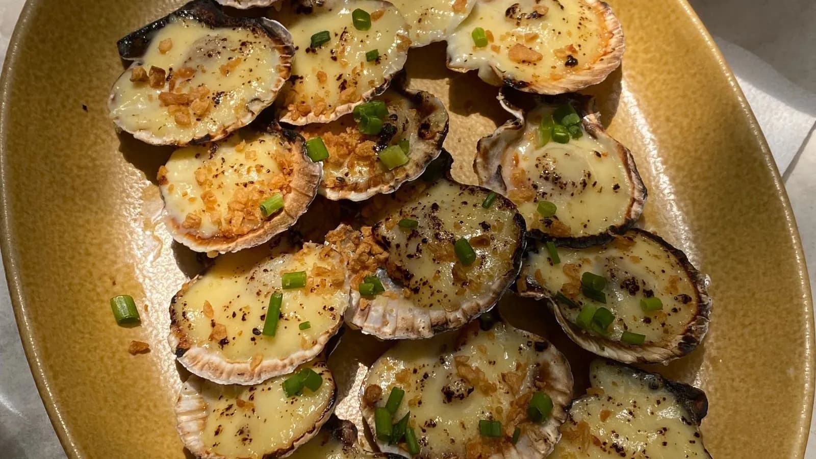 Vieiras gratinadas con queso fundido en sus conchas, más de una docena vistas desde arriba y solapadas sobre una fuente ovalada de cerámica ocre: cada una lleva una capa de queso de color crema, dorada y con motas chamuscadas, con ajo frito crujiente y cebolleta tierna picada por encima